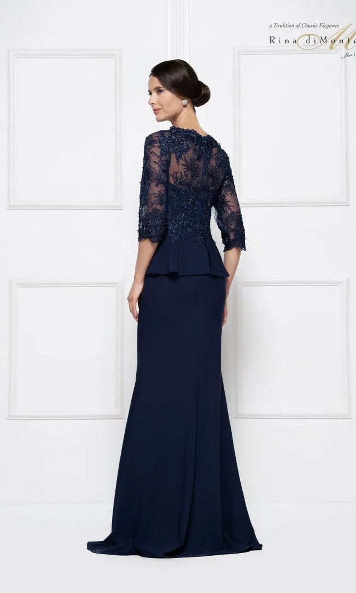 A woman stands with her back turned, wearing the Rina Di Montella RD2685 long formal dress in navy blue, featuring lace detailing on the bodice and elegant three-quarter embroidered sleeves.